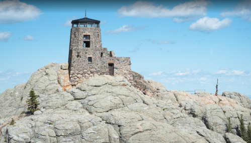 A stone lookout tower atop a rocky mountain, surrounded by blue sky and scattered clouds.