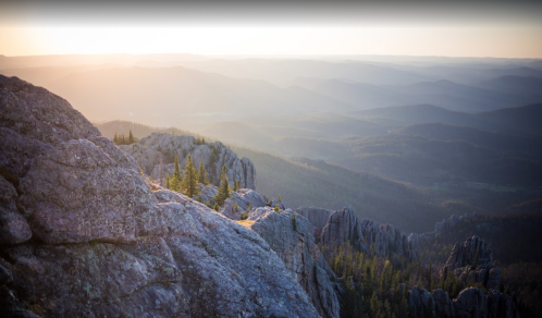 A panoramic view of rugged mountains at sunset, with rocky cliffs and distant hills bathed in warm light.