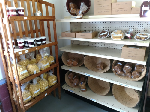 A wooden shelf displays jars and bags of pasta, while a second shelf holds baskets of baked goods and fruit.