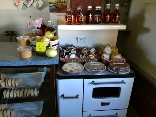 A countertop displays various baked goods, jars of syrup, and fresh apples, with a vintage stove in the background.