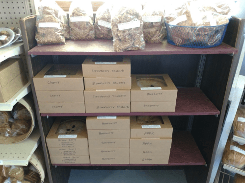 A wooden shelf displaying various pie boxes labeled with flavors like cherry, blueberry, and apple, alongside bags of granola.