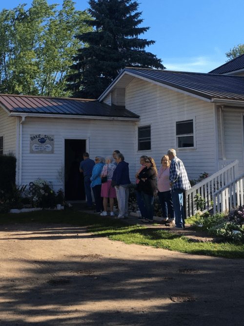 A line of people waiting outside a small bake shop on a sunny day, with trees in the background.