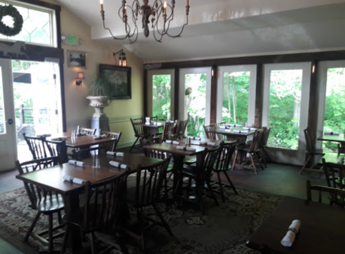 Cozy restaurant interior with wooden tables, chairs, and large windows overlooking greenery.