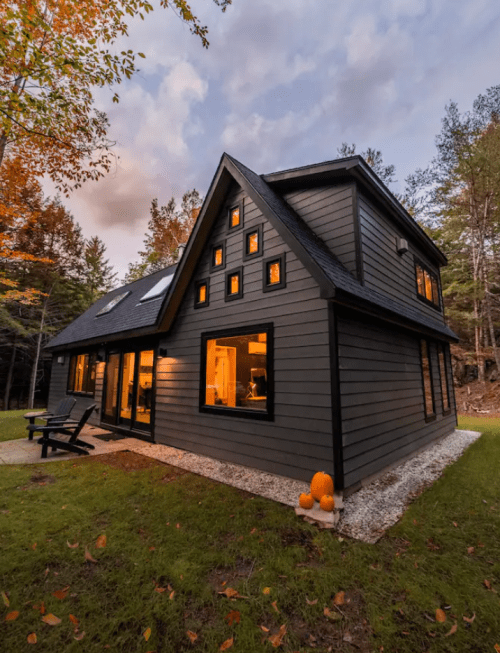 A modern dark gray house with large windows, surrounded by autumn foliage and pumpkins on the porch.