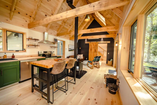 Cozy cabin interior with wooden beams, a kitchen island, and a wood stove, surrounded by large windows.