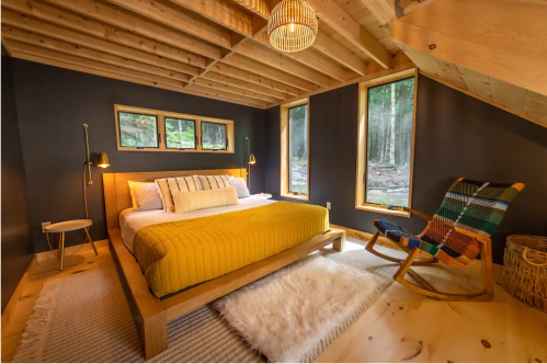 Cozy bedroom with a yellow bedspread, wooden accents, large windows, and a rocking chair, surrounded by nature.