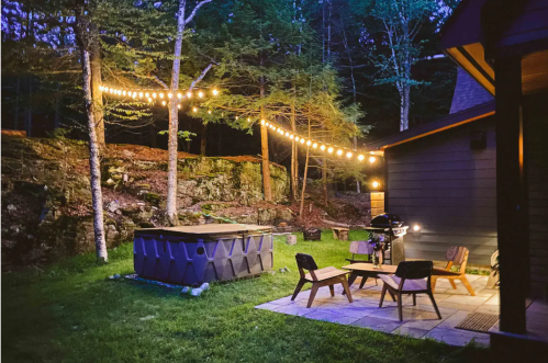 Cozy outdoor space with string lights, a hot tub, and seating area surrounded by trees at dusk.