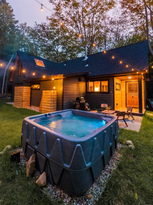 A cozy cabin with string lights and a hot tub in a serene outdoor setting surrounded by trees.