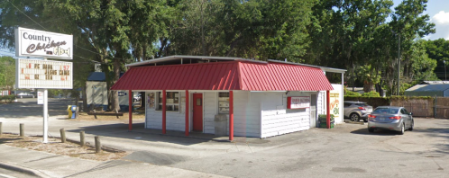 A small restaurant with a red metal roof and white walls, surrounded by trees and parked cars.