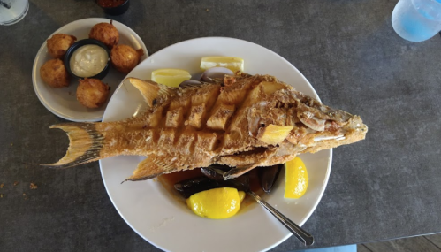 A whole fried fish garnished with lemon slices, served with hushpuppies and dipping sauce on the side.