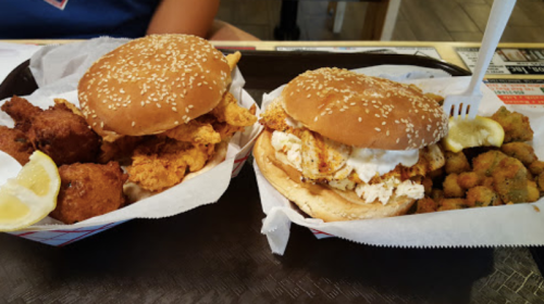 Two sesame seed burger buns filled with fried chicken and grilled chicken, served with fried okra and lemon wedges.