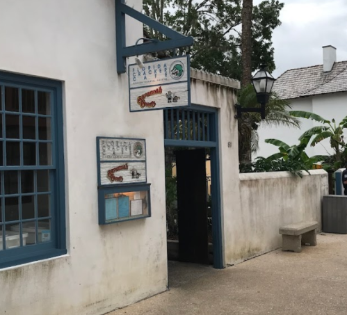 A quaint building entrance with blue accents and a sign, surrounded by greenery and a stone bench.
