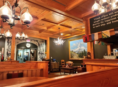 Cozy wooden interior of a restaurant with chandeliers, a large painting, and flags hanging on the wall.