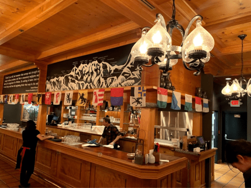 A cozy bar with wooden decor, featuring flags hanging above and staff preparing drinks in a warm atmosphere.
