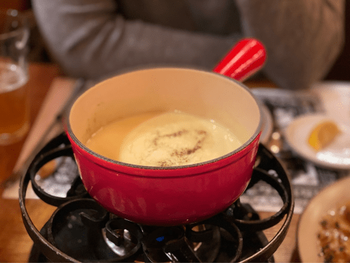 A red fondue pot filled with melted cheese, sitting on a black stand, with a person and a plate in the background.