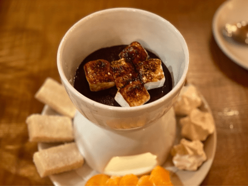 A bowl of chocolate fondue topped with toasted marshmallows, surrounded by fruit and bread on a wooden table.