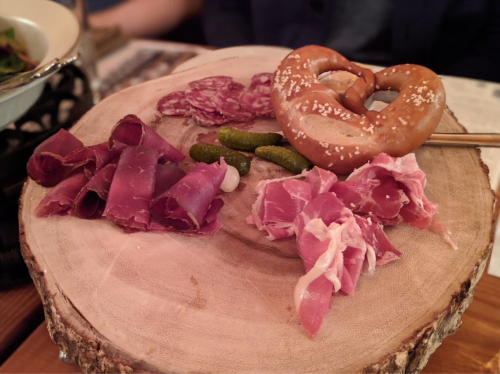 A wooden platter with assorted cured meats, pickles, and a pretzel.