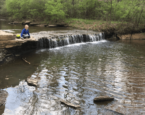 A child sits on a rock by a small waterfall, surrounded by greenery and a calm stream.