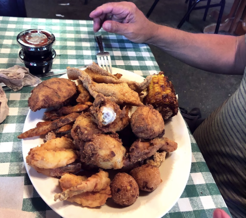 A large plate of assorted fried foods, including chicken, fish, corn, and hushpuppies, on a checkered tablecloth.
