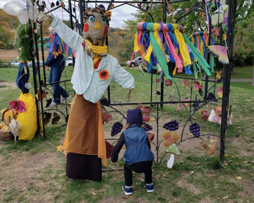 A colorful scarecrow with a sunflower stands in a park, while a child in a blue jacket plays nearby.