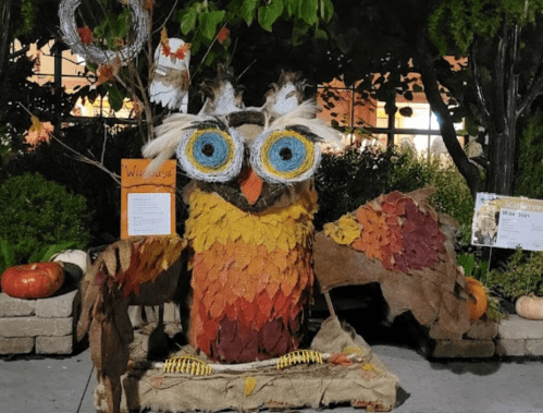 A large, colorful owl sculpture made of leaves and straw, surrounded by pumpkins and greenery in a festive setting.