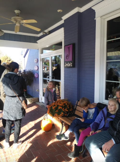 A group of people waits outside a shop with a purple exterior, featuring a bench and a pumpkin on the ground.