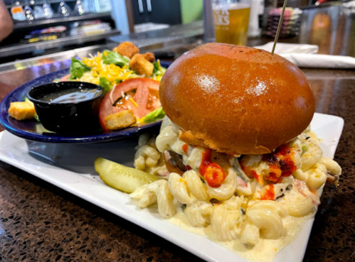 A plate featuring a burger topped with macaroni and cheese, a side salad, and a pickle on a textured surface.