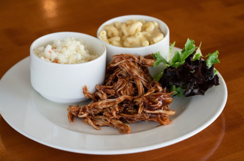 A plate with shredded meat, macaroni, coleslaw, and a side of mixed greens in white bowls.