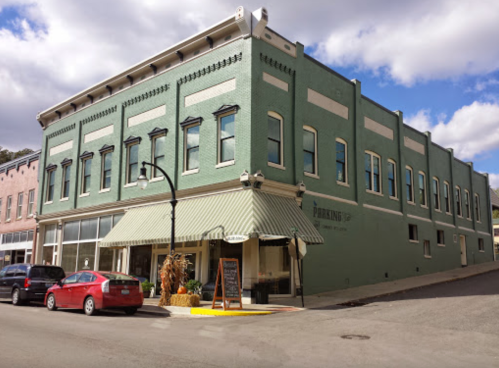 A green historic building on a corner, with a striped awning and parked cars nearby under a partly cloudy sky.