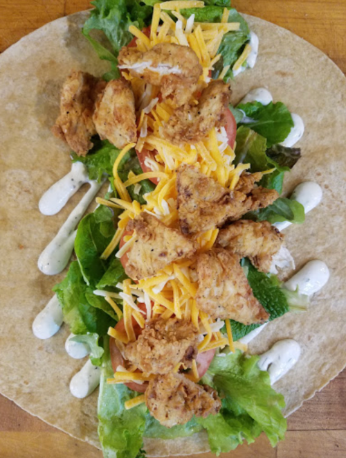 A tortilla wrap filled with lettuce, tomatoes, fried chicken, shredded cheese, and drizzled with dressing.