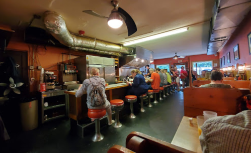 A cozy diner interior with patrons sitting at a counter and booths, featuring warm lighting and a vintage vibe.