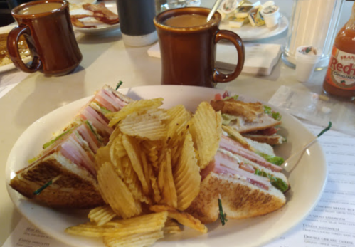 A plate of a club sandwich with layers of meat and lettuce, served with a side of potato chips and coffee mugs in the background.