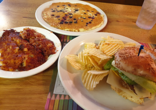 A plate with a sandwich, lettuce, cheese, and chips, alongside a pancake and hash browns on a wooden table.