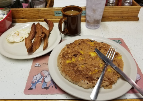 A plate with a large pancake, a side of potato wedges, a cup of coffee, and a glass of water on a diner table.