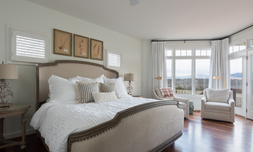 Bright, modern bedroom with a large bed, decorative pillows, and windows offering scenic views. Cozy armchair in the corner.