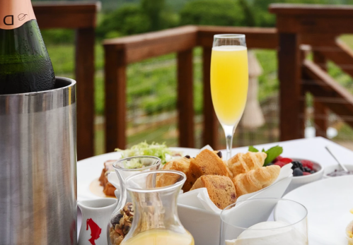 A table set with brunch items, including pastries, fruit, and a glass of mimosa, with a scenic vineyard in the background.