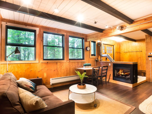 Cozy living room with a sofa, coffee table, and fireplace, surrounded by large windows and wooden accents.