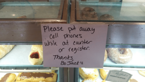 Sign in a bakery display case asking customers to put away cell phones at the counter or register.