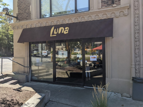 Exterior of a restaurant named "Luna," featuring large glass doors and a brown awning, surrounded by greenery.
