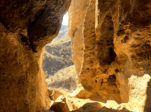 View from a rocky cave opening, revealing a sunlit landscape and distant hills.