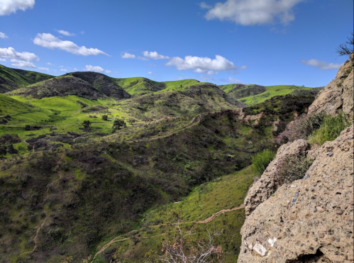 Lush green hills and valleys under a blue sky with scattered clouds, viewed from a rocky outcrop.