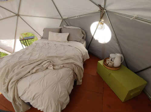 Cozy interior of a geodesic dome with a bed, soft blankets, and a small table with a pitcher and cups.