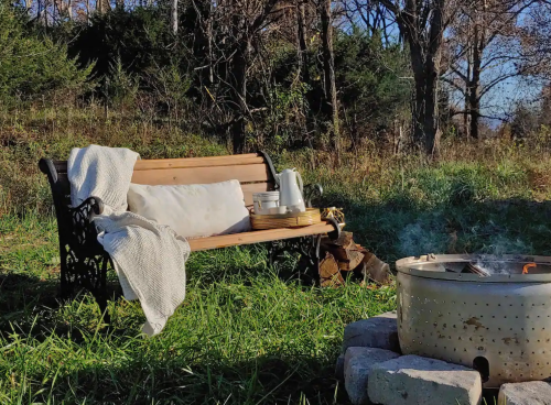 A cozy outdoor scene featuring a wooden bench with a blanket, next to a fire pit surrounded by greenery.