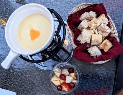 A bowl of cheese fondue with a heart-shaped topping, served with bread cubes and a side of mixed fruit.