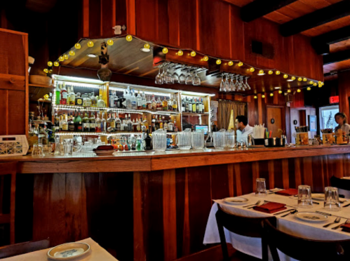 A cozy restaurant bar with wooden decor, shelves of liquor, and warm lighting, creating an inviting atmosphere.