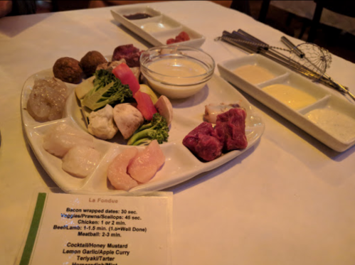 A platter of assorted meats and vegetables for fondue, accompanied by dipping sauces and a menu on the side.