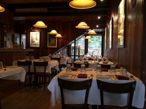 Cozy restaurant interior with wooden decor, tables set with white linens, and warm pendant lighting.
