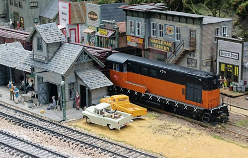 A model train scene featuring a black and orange locomotive, vintage cars, and small buildings with people.