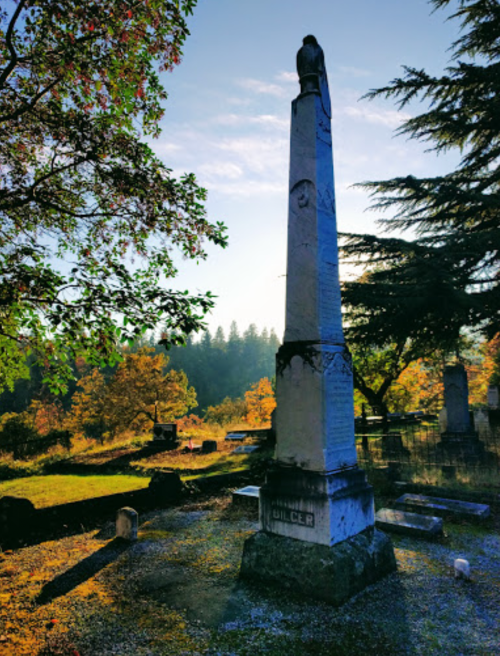 A tall, weathered monument in a serene cemetery, surrounded by autumn foliage and soft morning light.