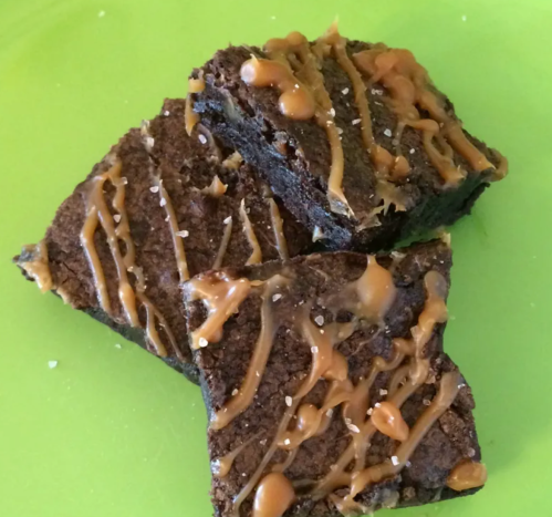 Three chocolate brownies drizzled with caramel sauce and sprinkled with sea salt, served on a green plate.